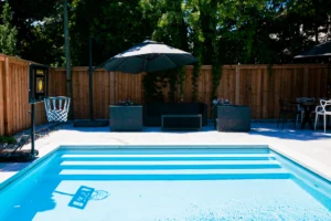 Shallow end of a custom inground pool showing wide tiled entry steps, a poolside basketball hoop, outdoor lounge seating under a patio umbrella, and a grey stone patio with a dining set in a Toronto backyard by Destination Pools and Landscaping