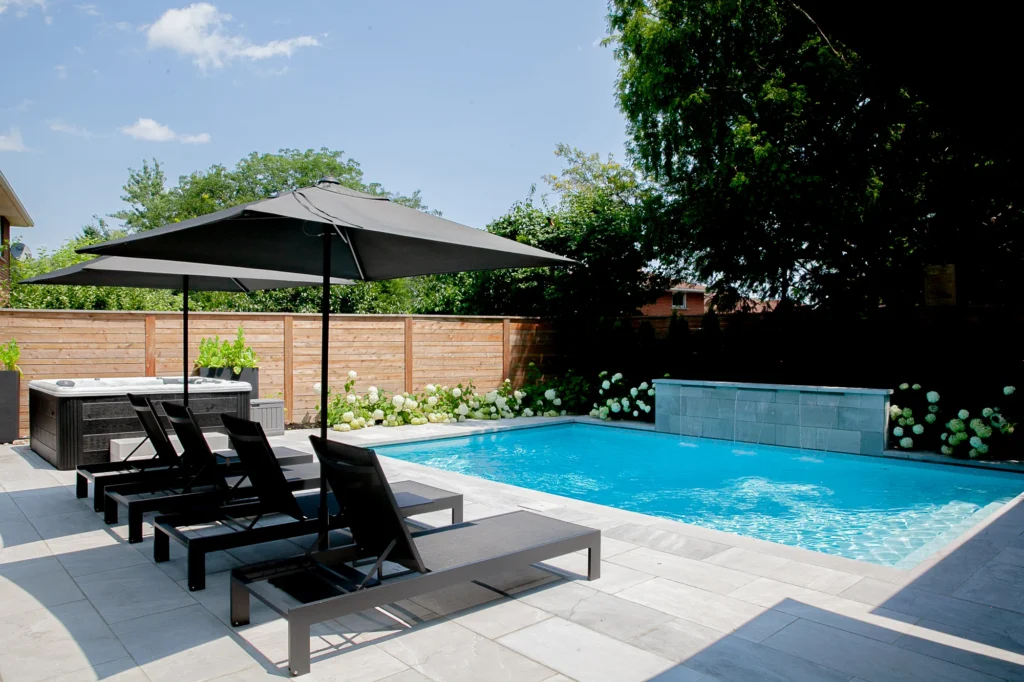 Inground pool with a hot tub, three black lounge chairs under a large patio umbrella, and a light grey porcelain stone deck, surrounded by cedar privacy fencing and white hydrangea shrubs in a Toronto backyard - Destination Pools and Landscaping