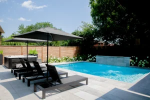 Inground pool with a hot tub, three black lounge chairs under a large patio umbrella, and a light grey porcelain stone deck, surrounded by cedar privacy fencing and white hydrangea shrubs in a Toronto backyard - Destination Pools and Landscaping