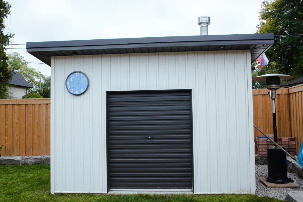 White pool equipment shed with a flat dark roof and dark roll-up door in a Mississauga backyard, housing pool mechanical systems installed by Destination Pools and Landscaping