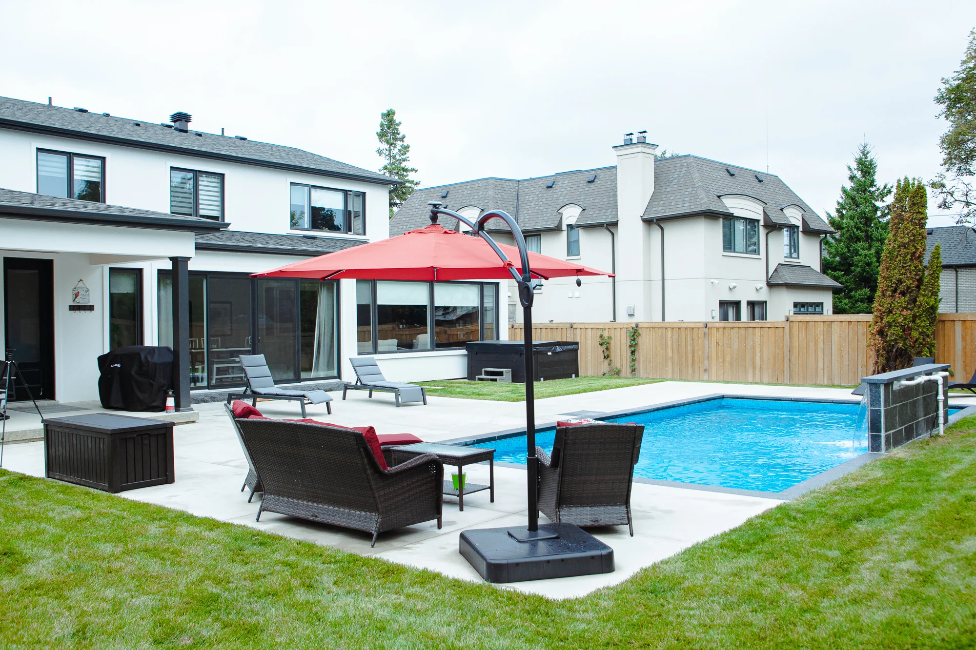 Wide-angle backyard view of a completed inground pool with grey stone patio, red outdoor sectional with cantilever umbrella, two dark chaise lounges, BBQ, and cedar privacy fence in Mississauga - by Destination Pools and Landscaping