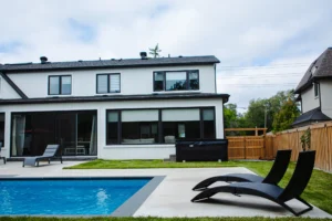 Rear of a white stucco Mississauga home with a rectangular inground pool, grey patio, two poolside chaise lounges, and a covered outdoor dining area - by Destination Pools and Landscaping