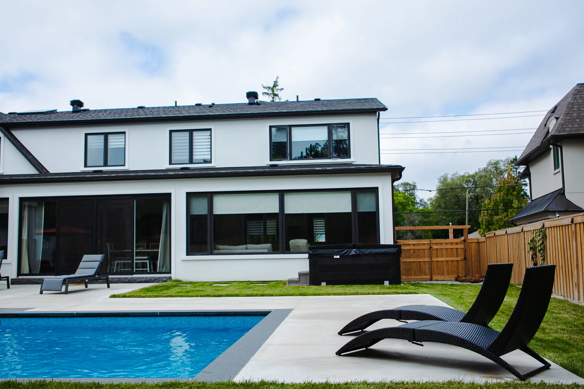 Rear of a white stucco Mississauga home with a rectangular inground pool, grey patio, two poolside chaise lounges, and a covered outdoor dining area - by Destination Pools and Landscaping