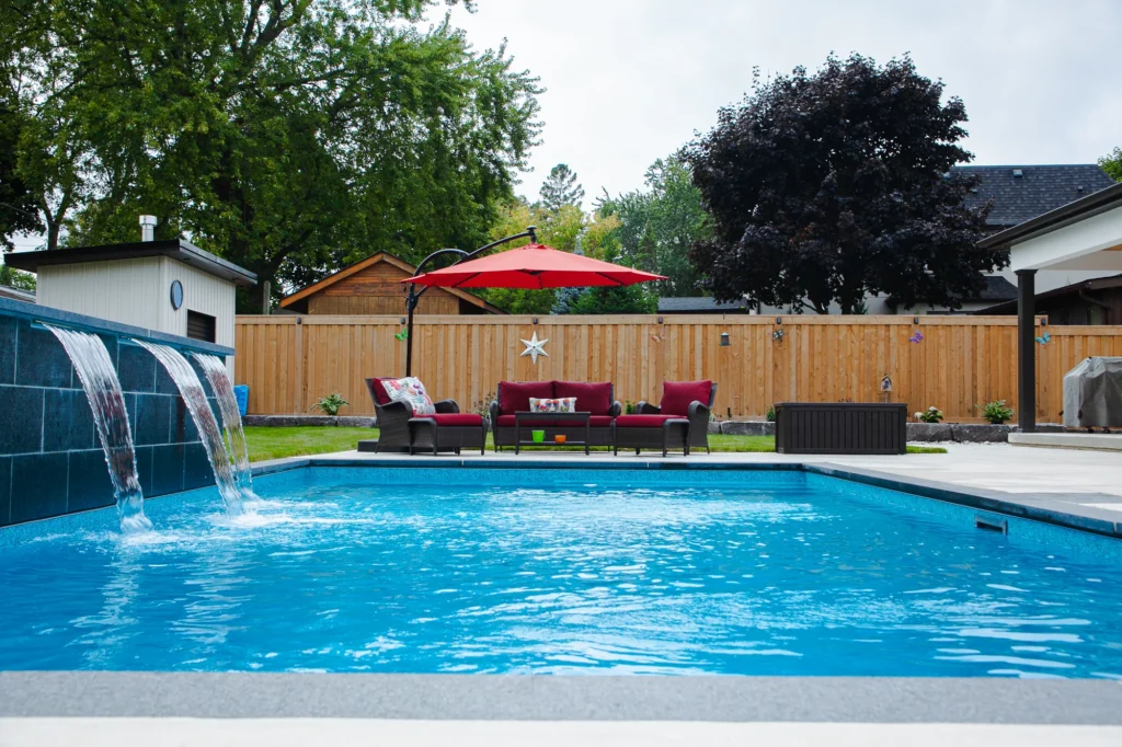 Rectangular inground pool with a three-sheet dark slate waterfall feature wall, blue pool water, red wicker sectional with umbrella, and cedar privacy fence in a Mississauga backyard - by Destination Pools and Landscaping