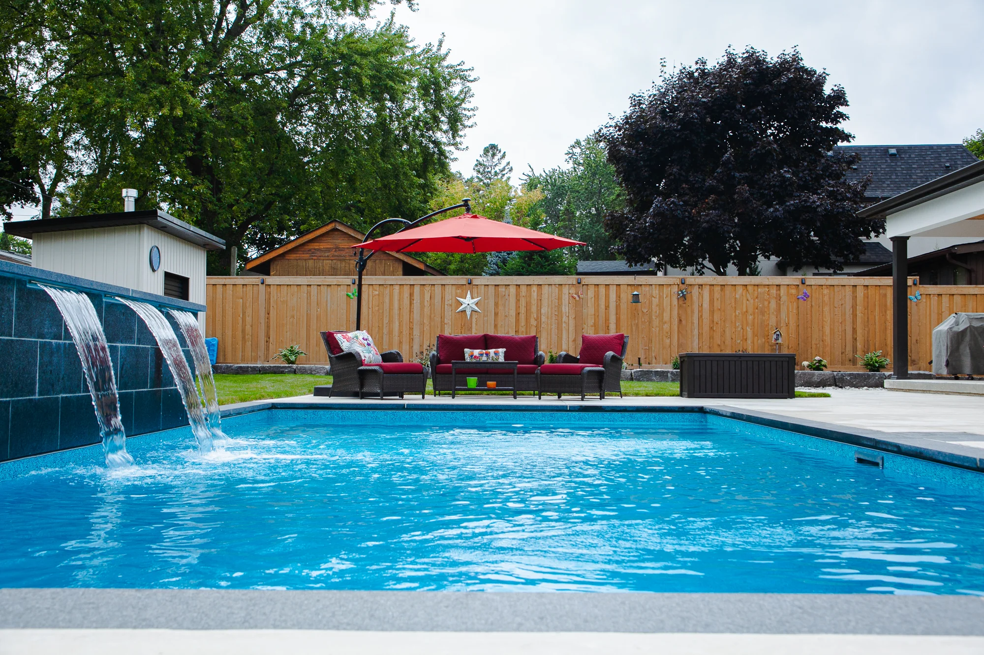 Rectangular inground pool with a three-sheet dark slate waterfall feature wall, blue pool water, red wicker sectional with umbrella, and cedar privacy fence in a Mississauga backyard - by Destination Pools and Landscaping