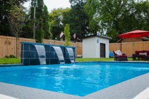 Inground pool with a three-sheet cascading dark slate waterfall feature, vibrant blue water, cedar fence, pool equipment shed, and red outdoor furniture in a Mississauga backyard - by Destination Pools and Landscaping