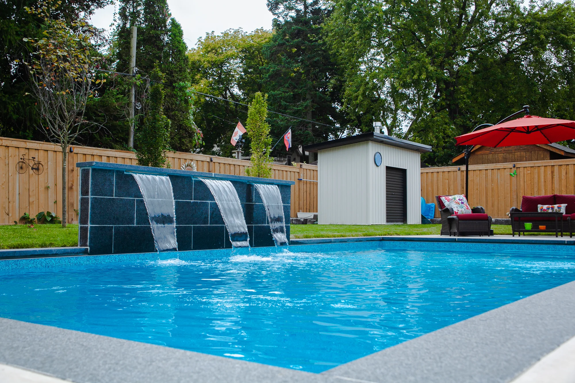 Inground pool with a three-sheet cascading dark slate waterfall feature, vibrant blue water, cedar fence, pool equipment shed, and red outdoor furniture in a Mississauga backyard - by Destination Pools and Landscaping