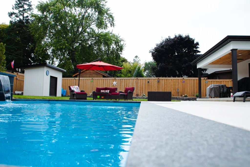 Ground-level view of a rectangular inground pool with grey coping, cascading waterfall feature, cedar fence, red outdoor furniture, and a covered pergola in a Mississauga backyard - by Destination Pools and Landscaping