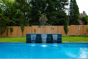 Closeup of a three-sheet dark slate waterfall feature wall cascading into a rectangular inground pool, with cedar privacy fence and greenery in the background - Mississauga pool by Destination Pools and Landscaping