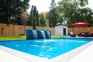 Full backyard view showing a rectangular inground pool with waterfall feature, grey coping, cedar fence, pool equipment shed, red cantilever umbrella, and a covered outdoor structure in Mississauga - by Destination Pools and Landscaping