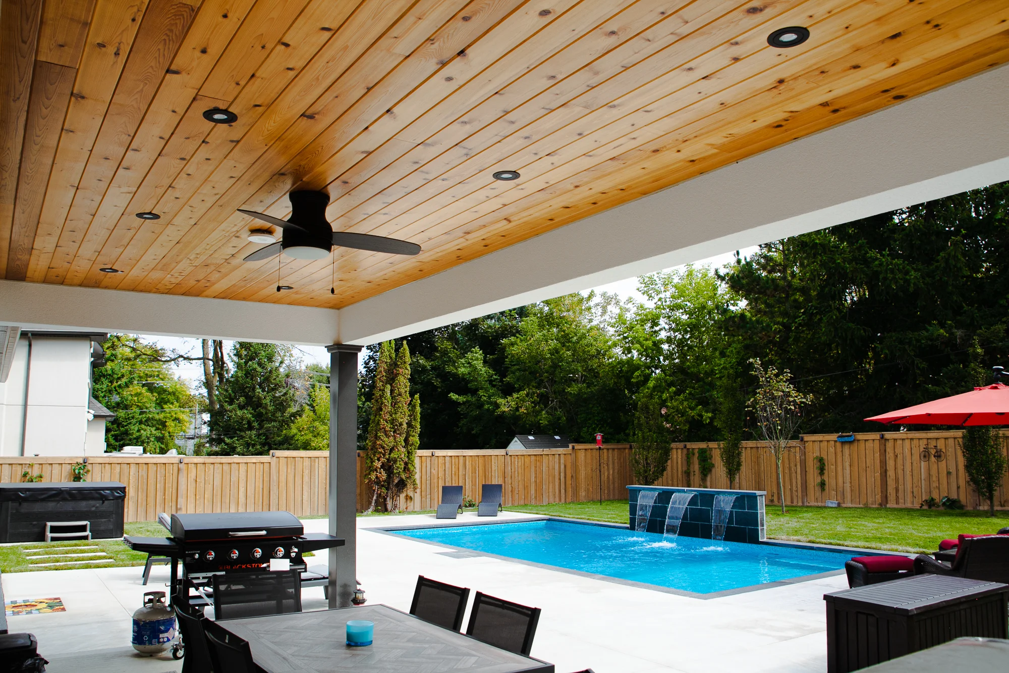 View from under a covered pergola with cedar wood ceiling, ceiling fan, and pot lights showing a BBQ, outdoor dining set, and rectangular inground pool with waterfall feature in the background - Mississauga backyard by Destination Pools and Landscaping