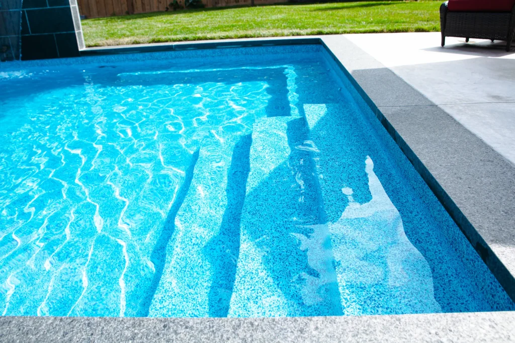 Close-up of the shallow end of a rectangular inground pool showing blue pebble aggregate vinyl liner, entry steps, and grey coping - Mississauga pool by Destination Pools and Landscaping