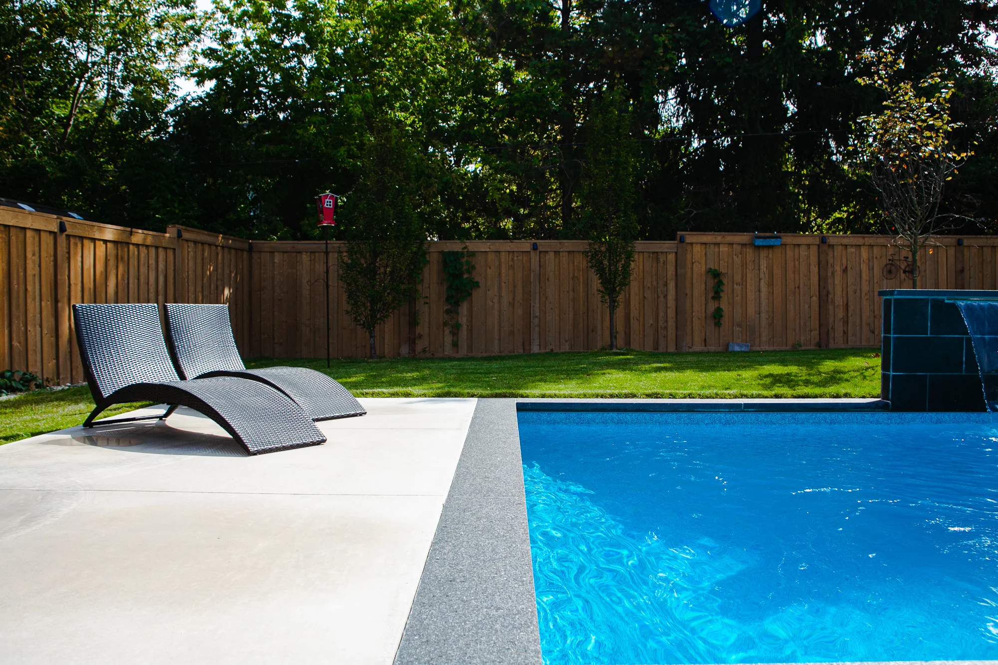Two dark wicker chaise lounges on a grey large-format patio beside a rectangular inground pool with a dark slate waterfall feature in a Mississauga backyard - by Destination Pools and Landscaping