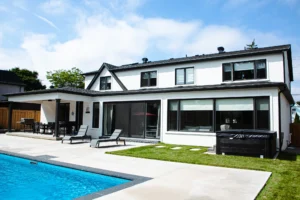 Sunny rear exterior of a white stucco Mississauga home with a grey patio, rectangular inground pool, covered pergola with dining set, chaise lounges, and a hot tub - by Destination Pools and Landscaping