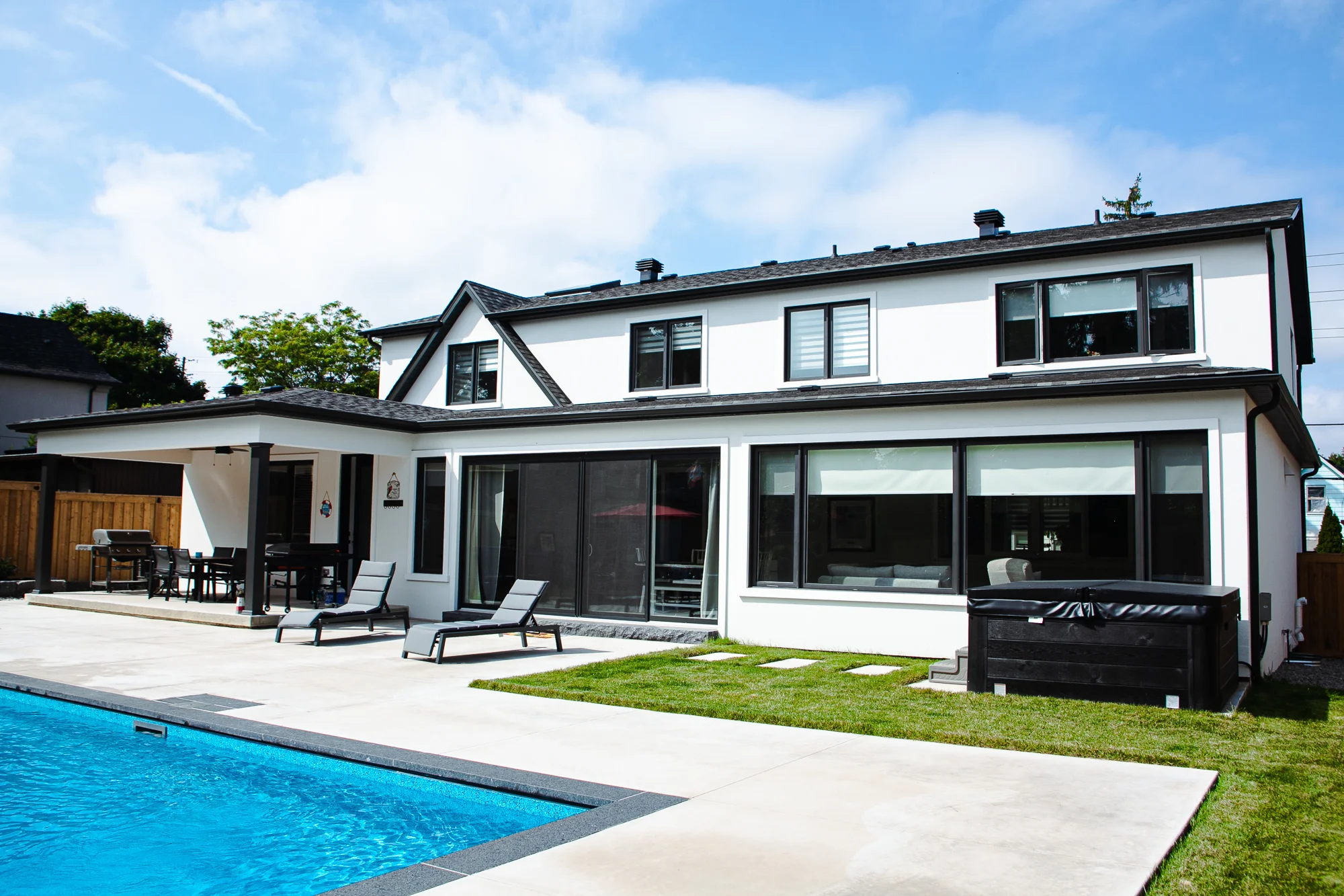 Sunny rear exterior of a white stucco Mississauga home with a grey patio, rectangular inground pool, covered pergola with dining set, chaise lounges, and a hot tub - by Destination Pools and Landscaping
