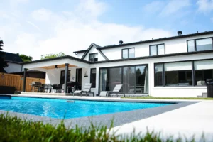Wide-angle rear view of a white stucco Mississauga home with a covered cedar-ceiling pergola, large rectangular inground pool, grey patio, and chaise lounges - by Destination Pools and Landscaping