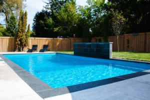 Rectangular inground pool with dark slate waterfall feature wall, grey coping, two dark chaise lounges, cedar fence, and pool equipment shed in a sunny Mississauga backyard - by Destination Pools and Landscaping