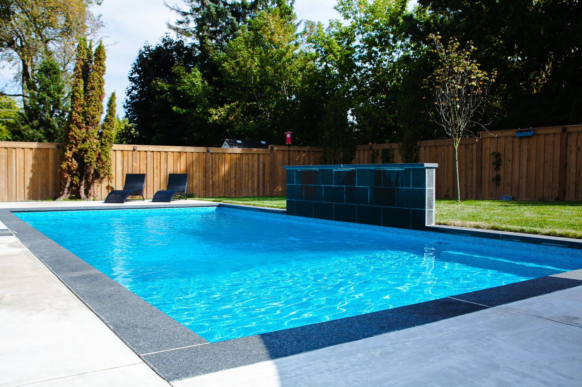 Rectangular inground pool with dark slate waterfall feature wall, grey coping, two dark chaise lounges, cedar fence, and pool equipment shed in a sunny Mississauga backyard - by Destination Pools and Landscaping