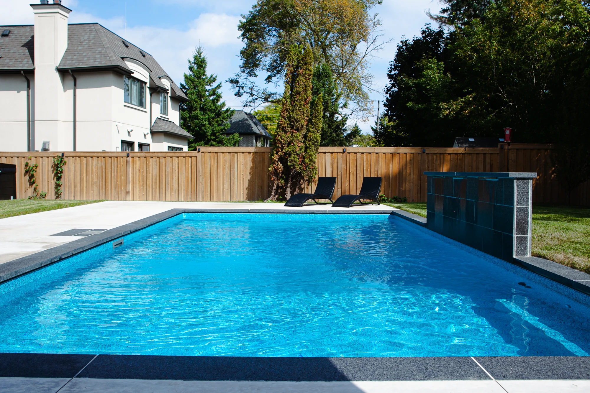 Full view of a rectangular inground pool with dark slate waterfall feature, grey coping, two dark chaise lounges, and cedar privacy fence in a Mississauga backyard - by Destination Pools and Landscaping