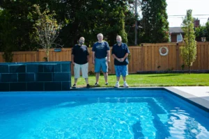 Three people standing beside an inground pool with a dark slate waterfall feature and cedar privacy fence in a Mississauga backyard - completed project by Destination Pools and Landscaping