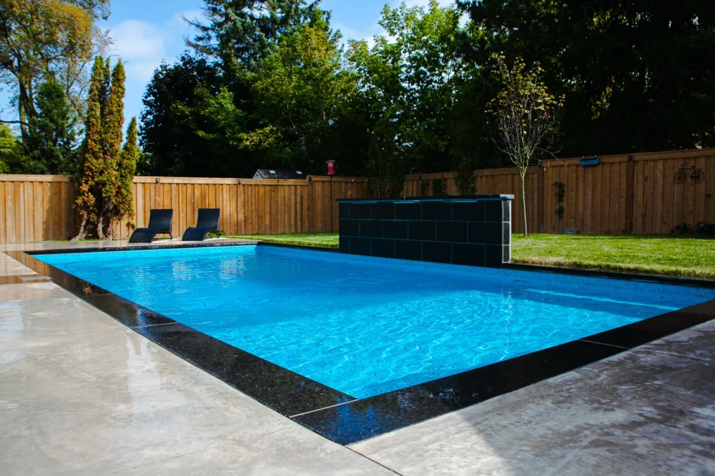 Rectangular inground pool with dark slate waterfall feature, grey large-format coping, two dark chaise lounges on the grass, and cedar fence with lush trees in Mississauga - by Destination Pools and Landscaping