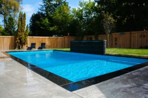 Rectangular inground pool with dark slate waterfall feature, grey large-format coping, two dark chaise lounges on the grass, and cedar fence with lush trees in Mississauga - by Destination Pools and Landscaping
