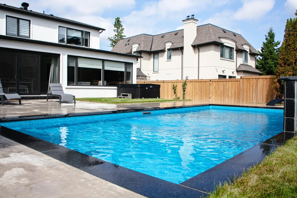 Full backyard view showing a rectangular inground pool, grey patio, dark chaise lounges, covered hot tub, cedar fence, and white stucco home with large windows in Mississauga - by Destination Pools and Landscaping