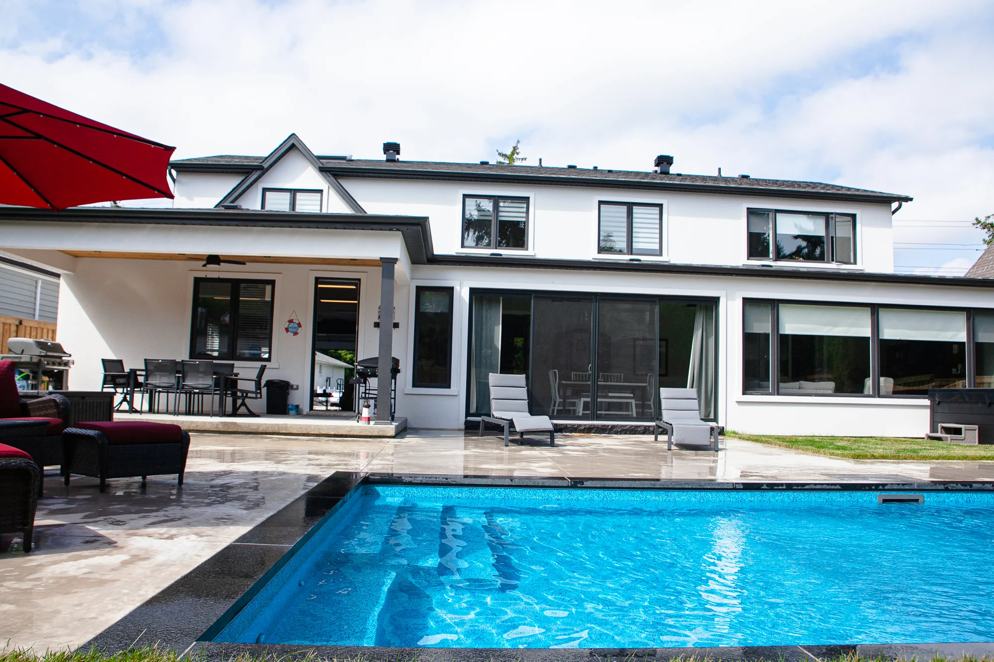 Wide rear view of a white stucco Mississauga home with a rectangular inground pool, grey patio, covered pergola with dining set, chaise lounges, and red umbrella - by Destination Pools and Landscaping