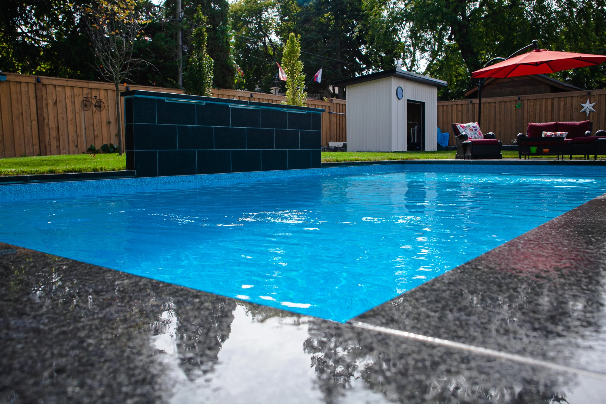Rainy-day view of a rectangular inground pool with dark slate waterfall feature, wet grey coping reflecting the surface, cedar fence, and pool equipment shed with red outdoor furniture in Mississauga - by Destination Pools and Landscaping
