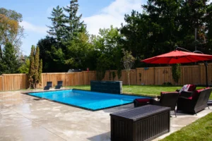 Full backyard view of a rectangular inground pool with grey large-format patio, dark slate waterfall feature, green lawn, red cantilever umbrella with wicker outdoor furniture, and cedar fence in Mississauga - by Destination Pools and Landscaping