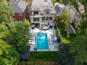 Aerial view of the pool, black cabana with outdoor shower, and rows of white cushioned loungers beneath a cantilever umbrella beside the lawn