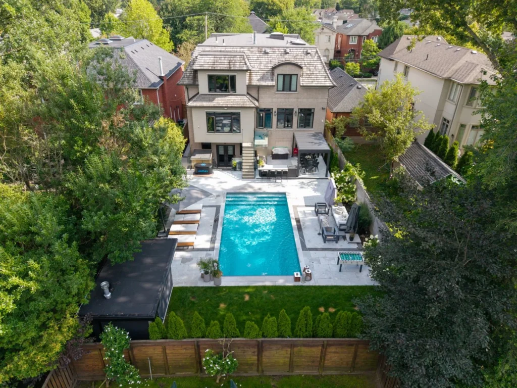 Aerial view of the full backyard showing the pool, patio furniture, black cabana, foosball table, and surrounding hedge and tree screening